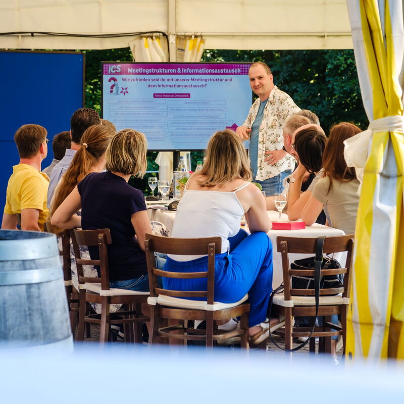 Teilnehmende sitzen an einem Tisch un verfolgen eine Präsentation